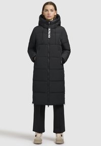 Zwarte pufferjack met een capuchon, voorzien van een rits aan de voorkant, elastische koordjes en een gewatteerd ontwerp. Gedragen over zwarte broeken en beigem schoenen.