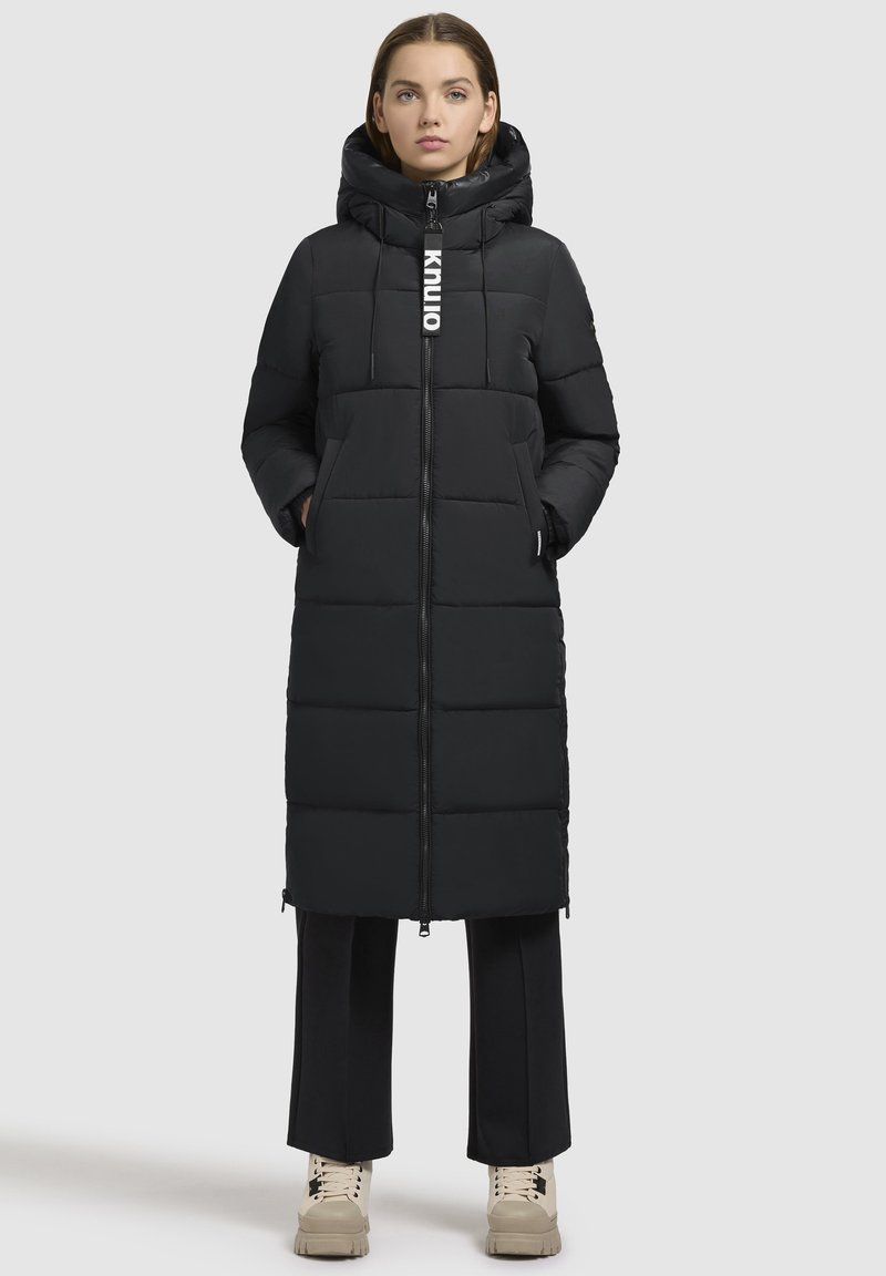 Zwarte pufferjack met een capuchon, voorzien van een rits aan de voorkant, elastische koordjes en een gewatteerd ontwerp. Gedragen over zwarte broeken en beigem schoenen.