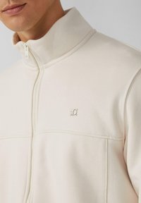 Hellbeige Zip-Jacke mit hohem Kragen, ausgestattet mit einem kleinen silbernen Logo auf der Brust und einem glatten, strukturierten Stoff.
