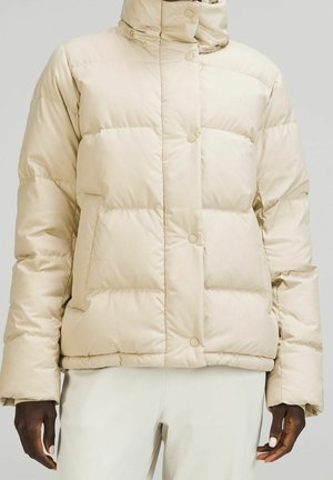 Veste matelassée beige avec boutons-pression et col montant portée par une personne à la peau foncée, associée à un pantalon blanc cassé.