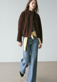 Braune Wildleder-Bomberjacke mit hohem Kragen, elastischem Bund, darüber ein gestreiftes Hemd und ein gebundenes gelbes Sweatshirt. Weit geschnittene blaue Jeans.