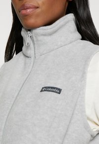 Harmaa fleece-liivi, jossa on korkea kaulus ja etuvetoketju. Rintamuksessa on musta Columbia-logo. Pehmeä pinta ja virtaviivainen muotoilu.
