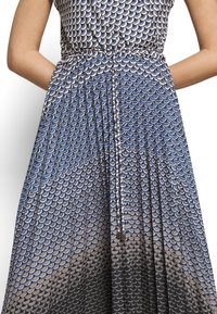 Kvinna som bär en blå och svart mönstrad klänning med en knuten midja, med händerna vilande bakom ryggen, mot en vit bakgrund.
