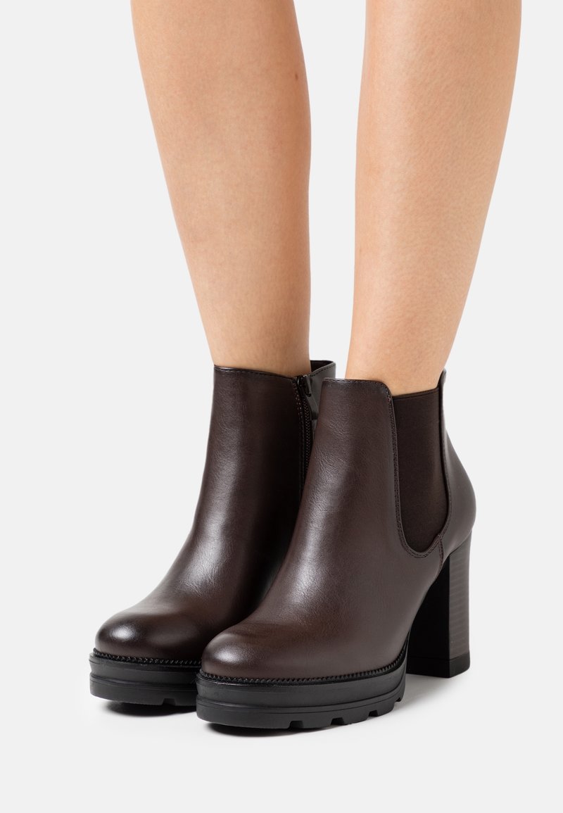 Bottines en cuir marron avec talons blocs et panneaux élastiques latéraux portées sur jambes nues sur fond blanc.