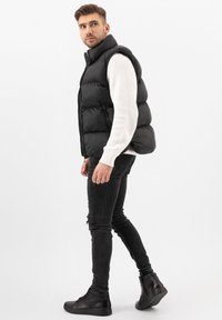 Černá puffa vesta s vysokým límcem a zipem, kombinovaná s bílým žebrovaným svetrem a černými úzkými džíny. Černé vysoké tenisky.