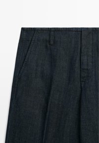 Tmavě modré džíny s postranními kapsami, poutky na opasek a záhyby viditelné v oblasti předního pasu na bílém pozadí.