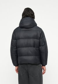 Chaqueta negra acolchada con capucha, que presenta secciones acolchadas horizontales y una textura suave. El diseño es de corte amplio y casual.