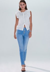 Fehér blúz fodros ujjakkal és részletes hímzéssel, világoskék szűk farmernadrággal párosítva, amelynek elején gombsor található.
