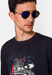 Czarne okulary przeciwsłoneczne z niebieskimi soczewkami, noszone przez mężczyznę w granatowym swetrze z grafiką nawiązującą do surfingu, przedstawiającą palmy i samochód.