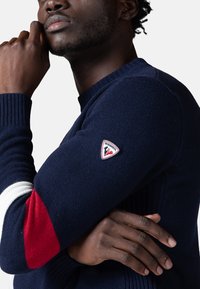 Maglione blu navy con scollo a girocollo, polsini a coste, una striscia rossa e bianca e una patch rotonda logata sulla manica sinistra.