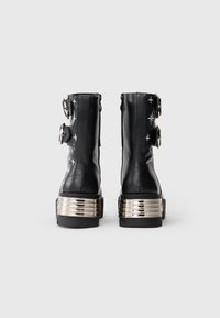 Botines de cuero negro hasta el tobillo con estrellas bordadas, dos hebillas plateadas cada uno y tacones metálicos plateados gruesos, mostrados desde la parte trasera.