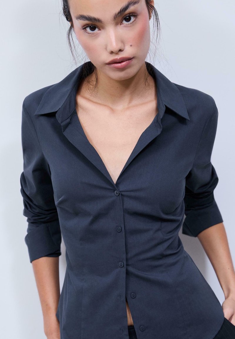 Dark blue button-up shirt with a collar, featuring long sleeves, a fitted design, and a smooth fabric texture. Buttons are visible down the front.