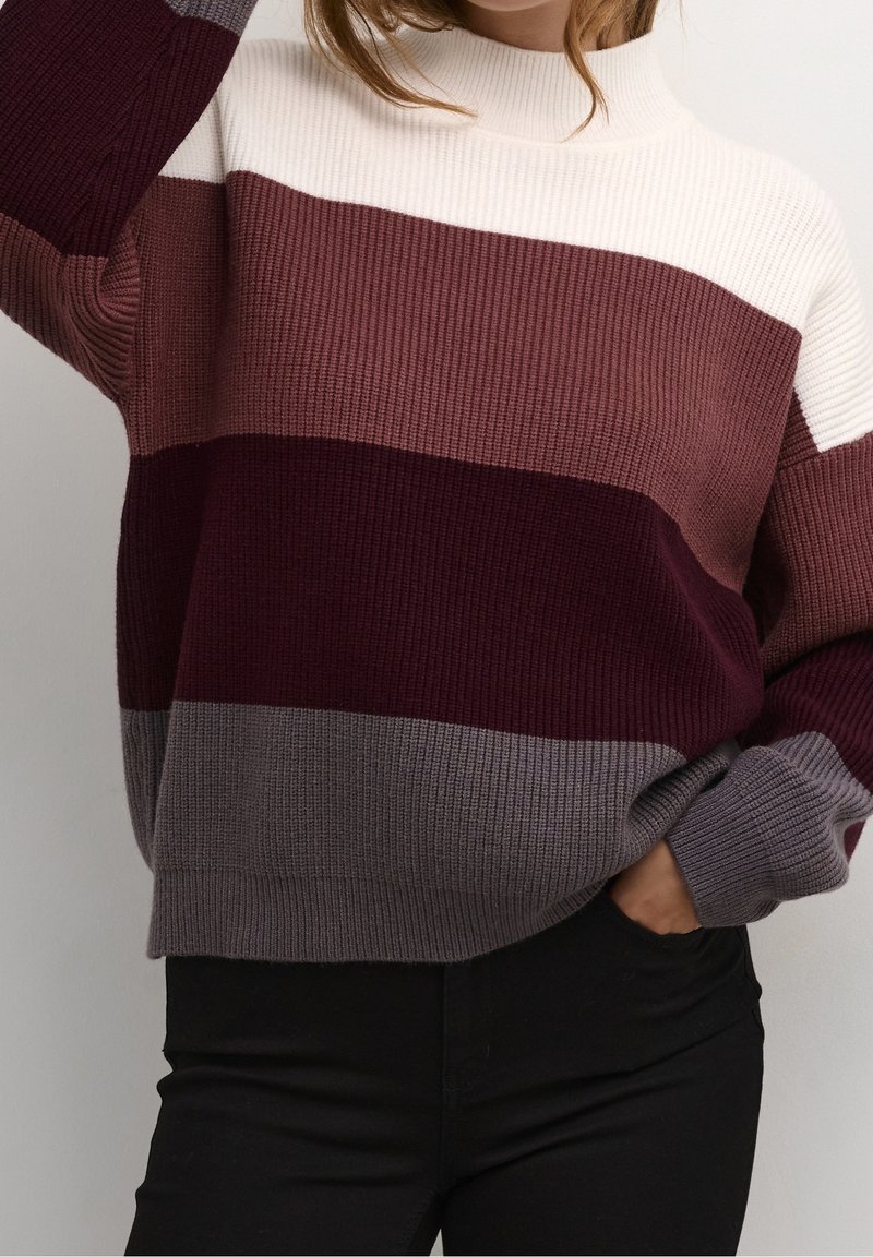 Femme portant un pull en maille côtelée avec de larges rayures horizontales blanches, mauves, bordeaux et grises, associé à un pantalon noir.