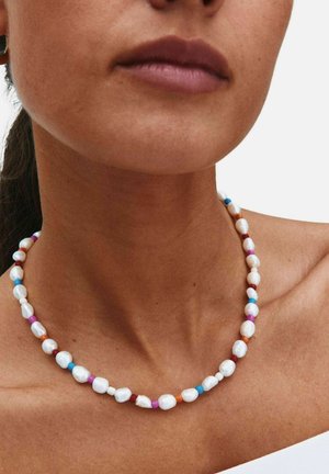 Ženska nosi ogrlico z nepravilnimi belimi biseri in majhnimi pisanimi perlicami v modri, roza, oranžni in rdeči barvi na svetlem tonu kože.