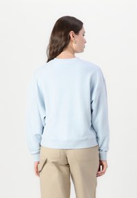 Felpa azzurra in morbido tessuto, con polsini e orlo a coste, dalla vestibilità comoda e design con scollo rotondo. Indossata con pantaloni beige.