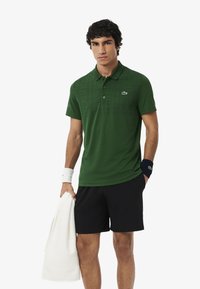 Polo a maniche corte di colore verde scuro con un motivo a quadretti, caratterizzato da un colletto, bottoni e il logo Lacoste. Indossato con pantaloni corti neri e un asciugamano bianco.