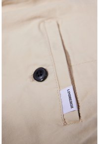 Gros plan sur un tissu beige avec un bouton noir et une petite étiquette blanche portant la mention « Lindbergh » cousue près de l'ouverture d'une poche.