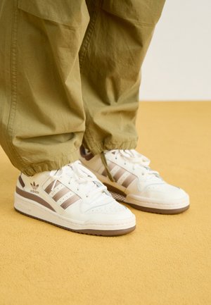 Białe i beżowe sneakersy Adidas noszone z luźnymi oliwkowozielonymi spodniami stojącymi na żółtej podłodze.