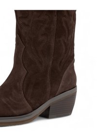 Bota vaquera de ante marrón con un diseño curvado, costuras decorativas y un tacón apilado. Presenta un acabado suave y texturizado.