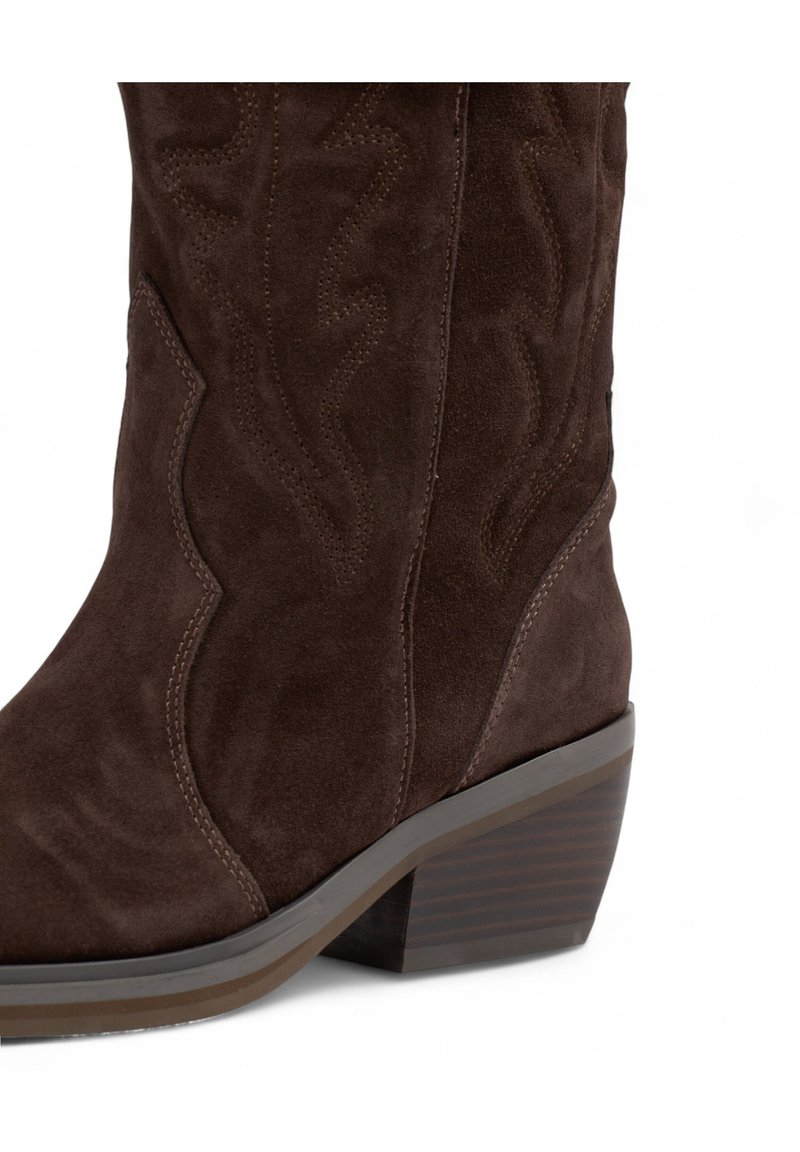 Bota vaquera de ante marrón con un diseño curvado, costuras decorativas y un tacón apilado. Presenta un acabado suave y texturizado.