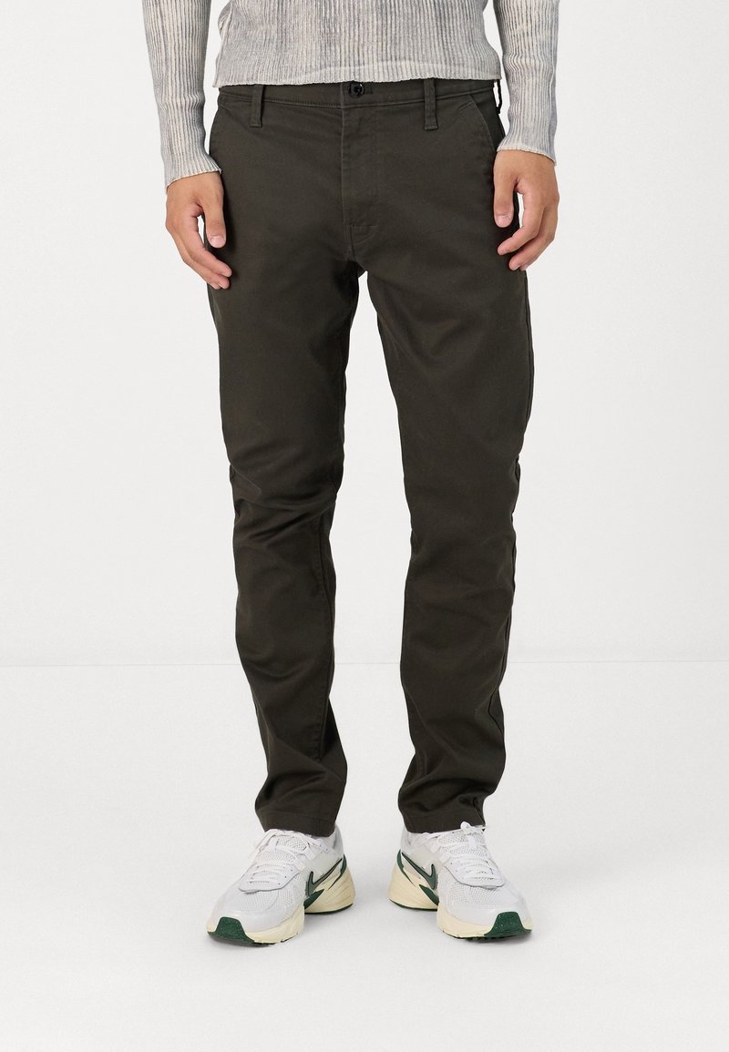 Homme portant un pantalon vert foncé, des baskets blanches avec des touches vertes et un t-shirt gris clair à manches longues côtelé, debout devant un fond blanc.
