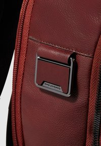 Sac à dos en cuir marron avec une finition texturée, doté d'un accent métallique argenté et d'une ouverture zippée. Des coutures détaillées sont visibles.