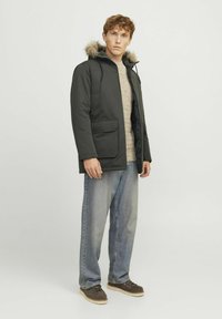 Donkergroene parka met een met bont gevoerde capuchon, voorzakken en elastische manchetten, gecombineerd met lichtblauwe jeans en bruine schoenen.