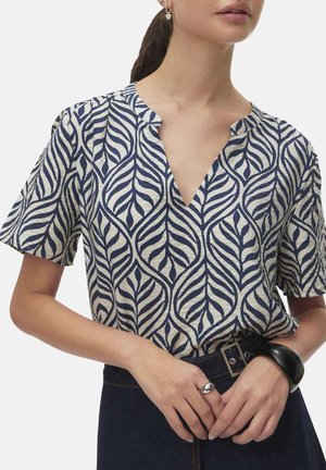 Femme portant un chemisier à manches courtes avec un motif de feuilles bleu et blanc, assorti d'une ceinture foncée et de bijoux aux mains et aux oreilles.