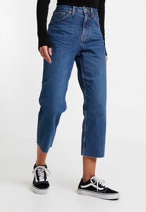 Person wearing high-waisted, cropped blue jeans and black-and-white lace-up sneakers, standing with one leg slightly forward.