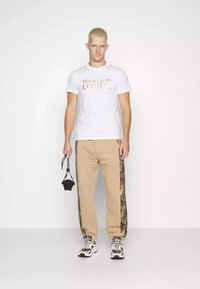 T-shirt bianca con "VERSACE JEANS COUTURE" in oro, pantaloni felpati beige con motivi laterali neri e sneakers chunky multicolori.