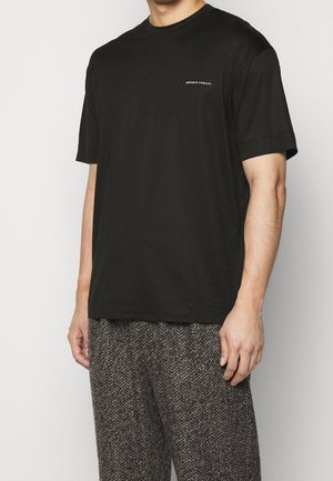 Mand iført en ensfarvet sort t-shirt med korte ærmer og små hvide tekst på brystet samt teksturerede grå mønstrede bukser.