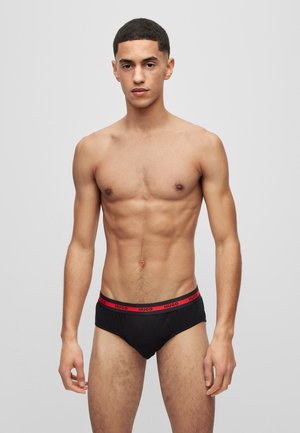 Schwarze Baumwollslips mit einem roten "HUGO"-Gummibund. Das Design umfasst einen tief sitzenden Schnitt und eine glatte Textur. Modell zeigt eine Vorderansicht.