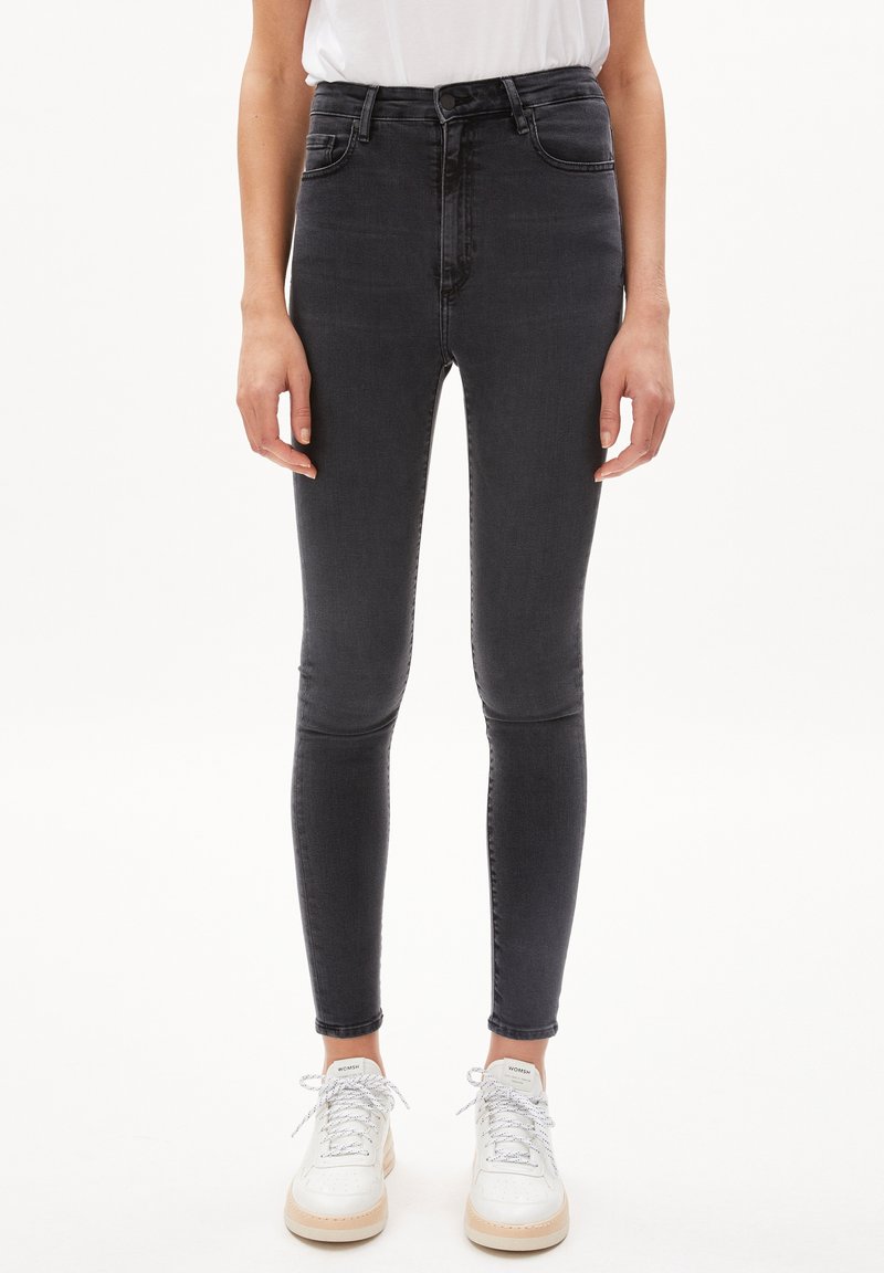 Černé skinny džíny s vysokým pasem vyrobené z denimu, s hladkou texturou a klasickým 5-kapanovým designem, doplněné bílými teniskami.