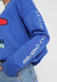Blue sweatshirt with red and green graphics, white text on the sleeve, ribbed cuffs, and a relaxed fit. Text reads “SAILING GEAR 88/88.”