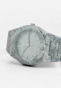 Reloj redondo de camuflaje gris en policarbonato con una correa texturizada, esfera minimalista y marcadores de hora plateados. Cuenta con una corona pulida.