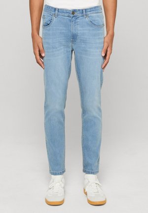 Personne portant un jean slim bleu clair et des baskets blanches à semelles en caoutchouc, debout devant un fond gris uni.