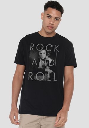 Young man wearing black t-shirt with vintage photo of a musician holding a guitar and text "ROCK AND ROLL" on front.