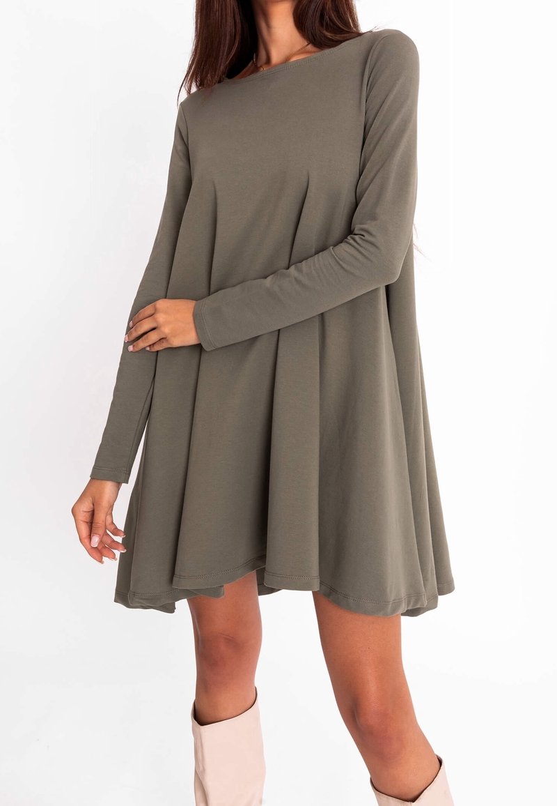 Femme portant une robe ample à manches longues de couleur vert olive avec des plis, associée à des bottes hautes beige clair, debout devant un fond blanc.