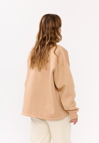 Felpa beige oversize con spalle scese, texture morbida e orlo arrotondato. Indossata sopra pantaloni chiari, che mostrano un taglio e un design casual.