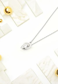 Silverhalsband med en tvinnad hänge som har en rad klara stenar, placerad mot en ljus marmorliknande bakgrund med guldfärgade accenter.