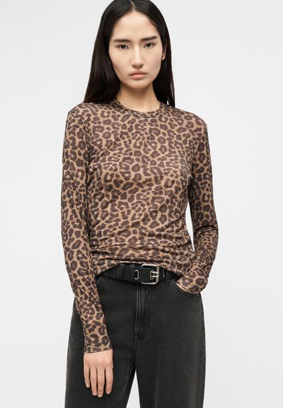 Jonge vrouw met lang donker haar, gekleed in een bruin shirt met luipaardprint en lange mouwen, ingestopt in zwarte jeans met een zwarte riem.