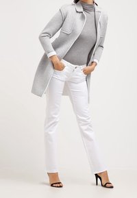 Abrigo largo gris sobre un suéter de cuello alto gris, combinado con jeans blancos ajustados y tacones de aguja negros. Tejido suave con corte a medida.