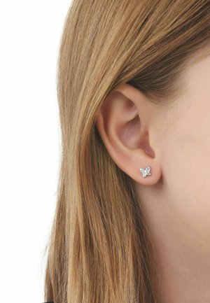 Primer plano de la oreja de una persona con cabello liso de color castaño claro que lleva un pequeño pendiente de botón plateado con forma de mariposa.