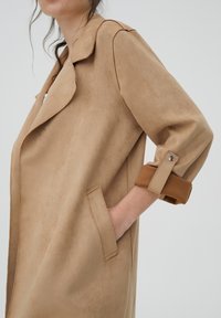 Femme portant un manteau en suède beige aux manches retroussées, main dans la poche, devant un fond uni gris clair.