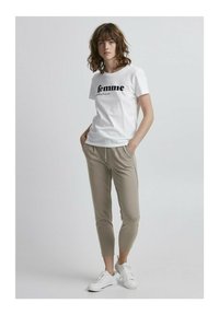 Hvit bomulls T-skjorte med "femme" trykt i svart, kombinert med beige tapered bukser og hvite sneakers. Modellen står med hendene i lommene.