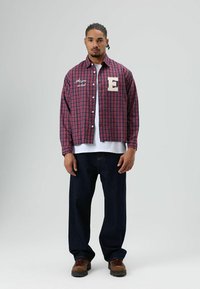 Rood-blauw geruit overhemd met witte knopen, voorzien van een geborduurde "E" en tekst aan de linkerkant, gecombineerd met donker denim broek en bruine schoenen.