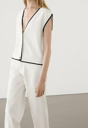 Personne portant un gilet blanc sans manches à col en V avec bordure noire et un pantalon blanc à jambes larges assorti, debout devant un fond uni.