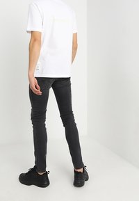 Camiseta blanca de manga corta con el texto "AESTHETIC" en amarillo, combinada con jeans negros ajustados y zapatillas negras, contra un fondo neutro.