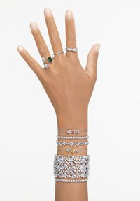 Linke Hand mit fünf silbernen Ringen mit Edelsteinen und fünf silbernen Armbändern, die klare, grüne, rosa und gelbe Steine auf weißem Hintergrund zeigen.