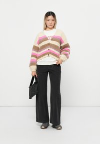Overmaat, gestreepte cardigan in beige, bruin, roze en wit met knopen. Gecombineerd met een witte sweatshirt en zwarte wijde broek.